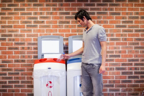 A young man with dark hair, wearing a light grey long-sleeve shirt and grey trousers, is standing outdoors against a red brick wall. He is holding a clear plastic bottle in his right hand and is about to dispose of it in a white recycling bin situated next to a similar bin, both with red and blue recycling symbols. The bins are positioned on a paved surface, likely part of a residential or commercial area in Wandsworth. The scene is well-lit, with natural daylight illuminating the area, emphasizing the textures and colors of the bricks, the man's clothing, and the bin materials. This setup reflects typical rubbish and recycling collection arrangements, aligning with services offered by Wandsworth House Clearance, especially in relation to waste segregation and disposal services for household or commercial waste in the local area.