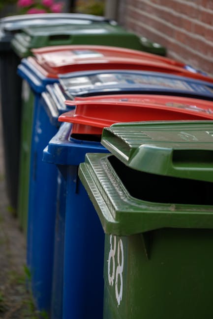 A row of large, rectangular wheelie bins with closed lids, positioned on a pavement alongside a red brick wall. The bins are primarily green, blue, and red, each with textured, ribbed plastic surfaces